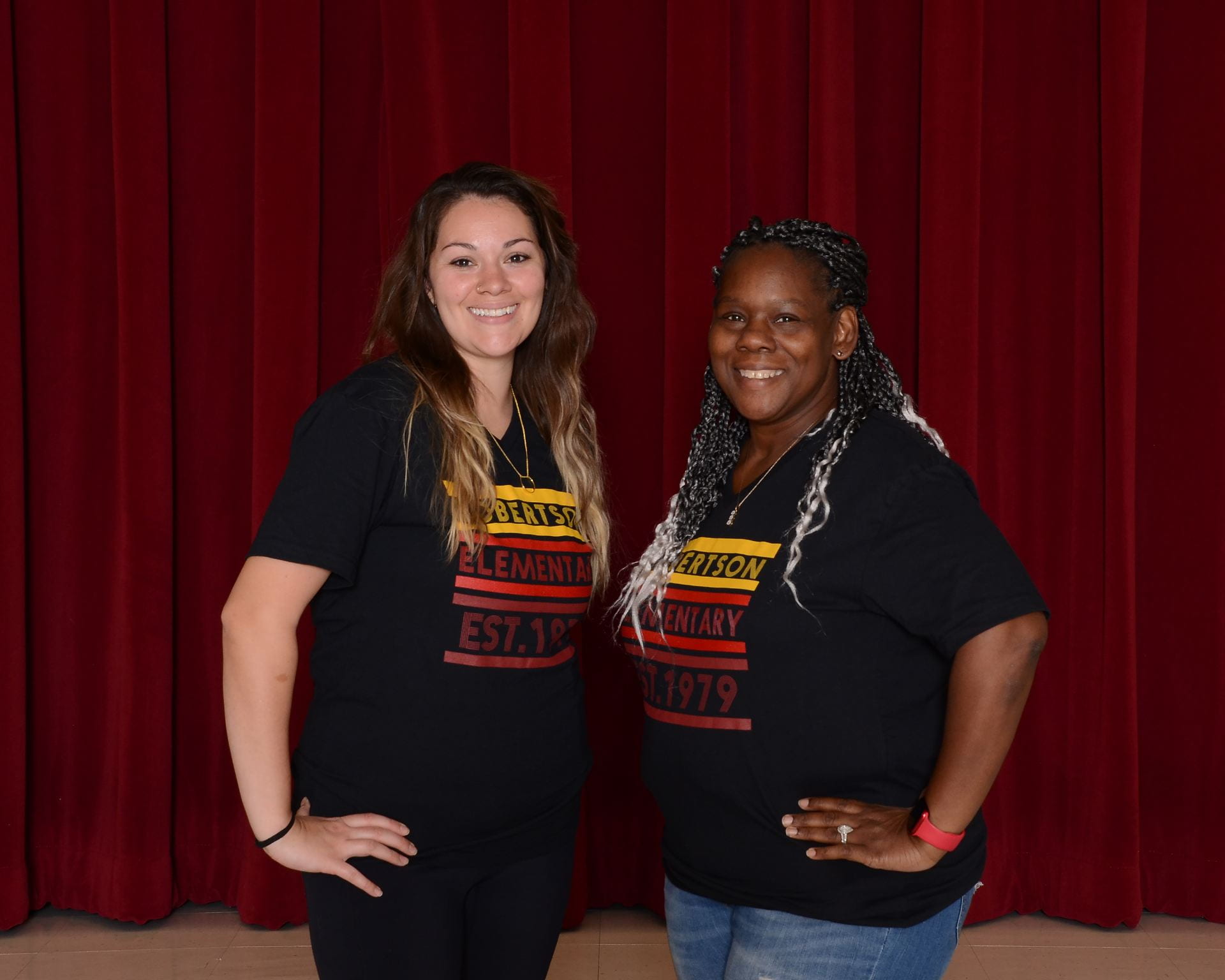 Two staff members wearing matching school shirts, stand in front of a dark background