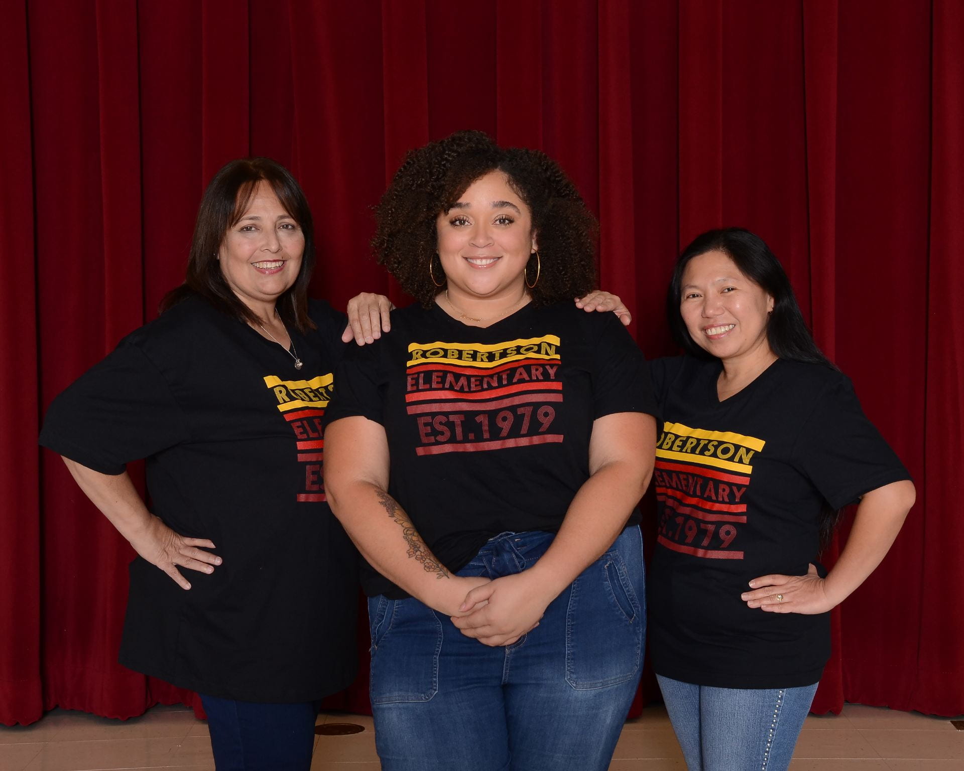 (3) First grade team teachers wearing school shirts, standing in front of a maroon stage curtain