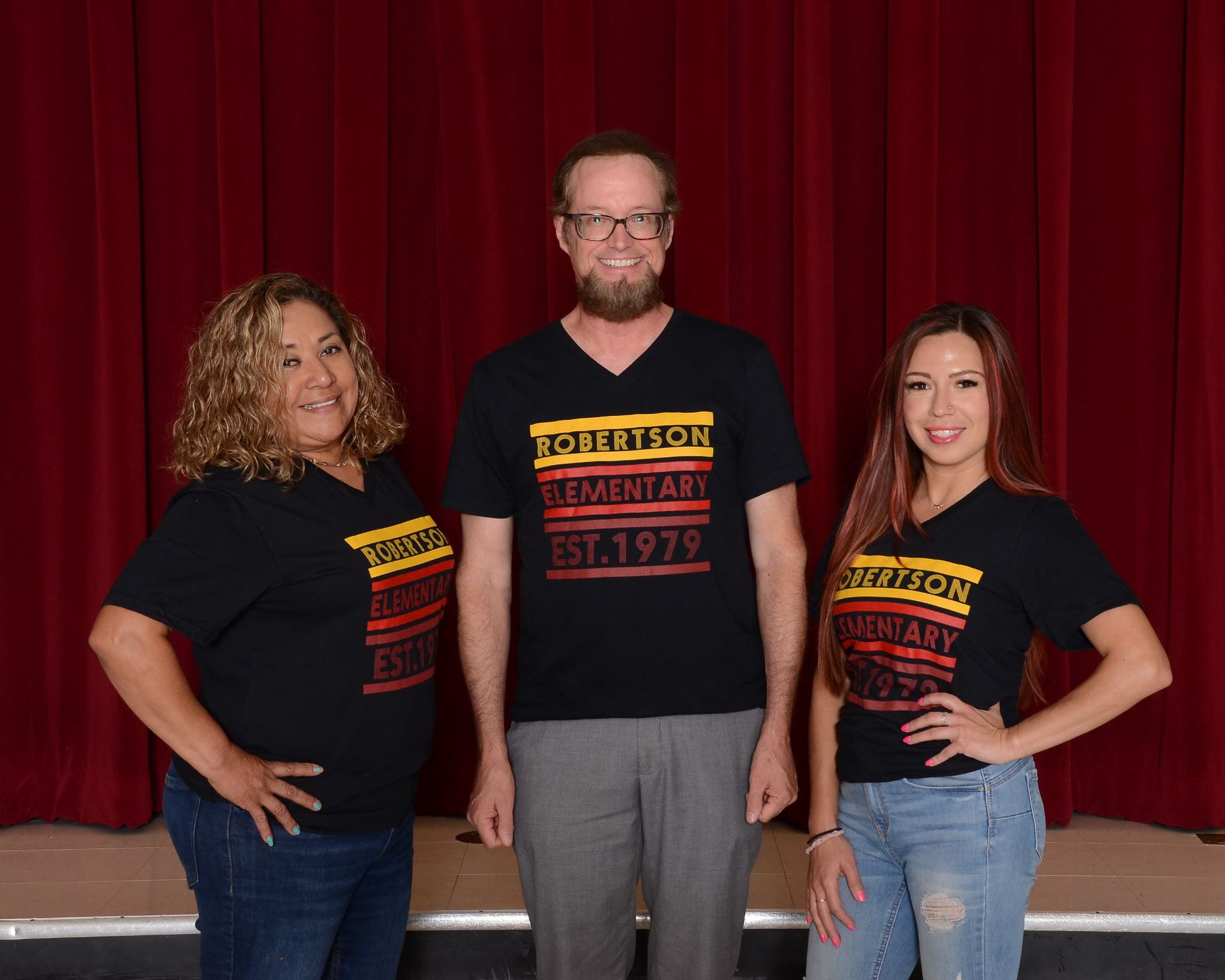 (3) Fourth grade team teachers wearing school shirts, standing in front of a maroon stage curtain