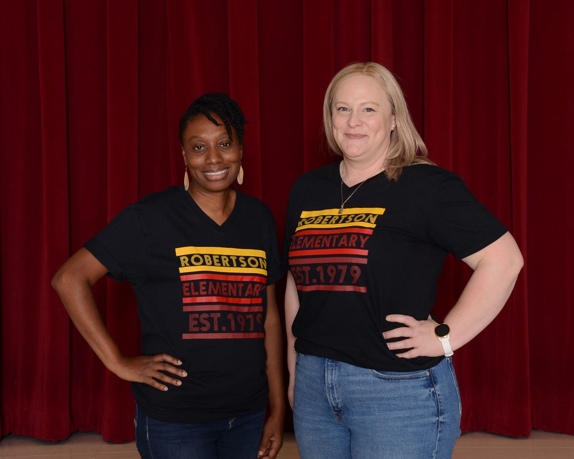 Two staff members wearing matching school shirts, stand in front of a dark background