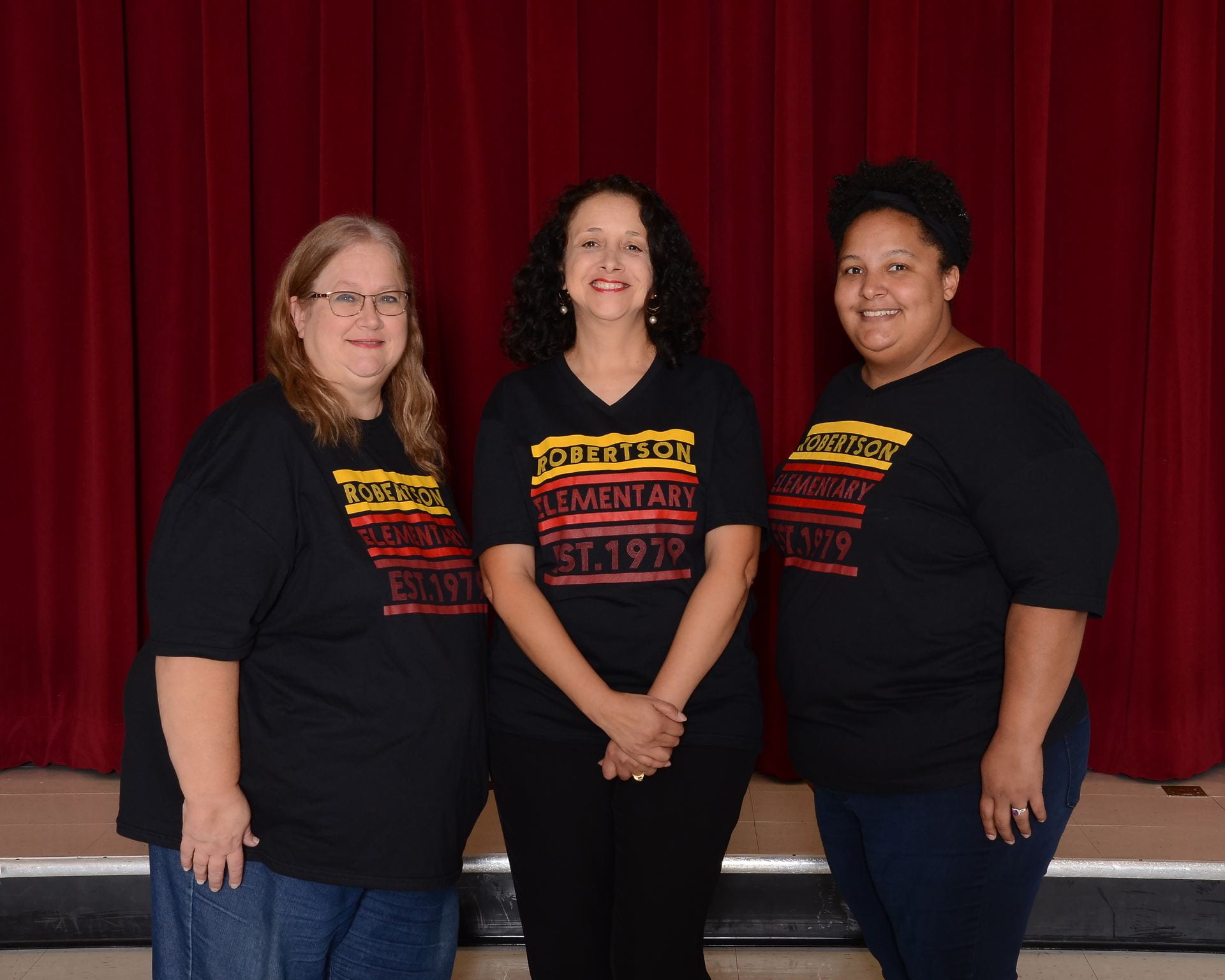 (3) Second grade team teachers wearing school shirts, standing in front of a maroon stage curtain
