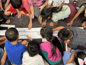 large group of second grade students kneel around a white paper with coding lines drawn on it, watching two robots follow the lines